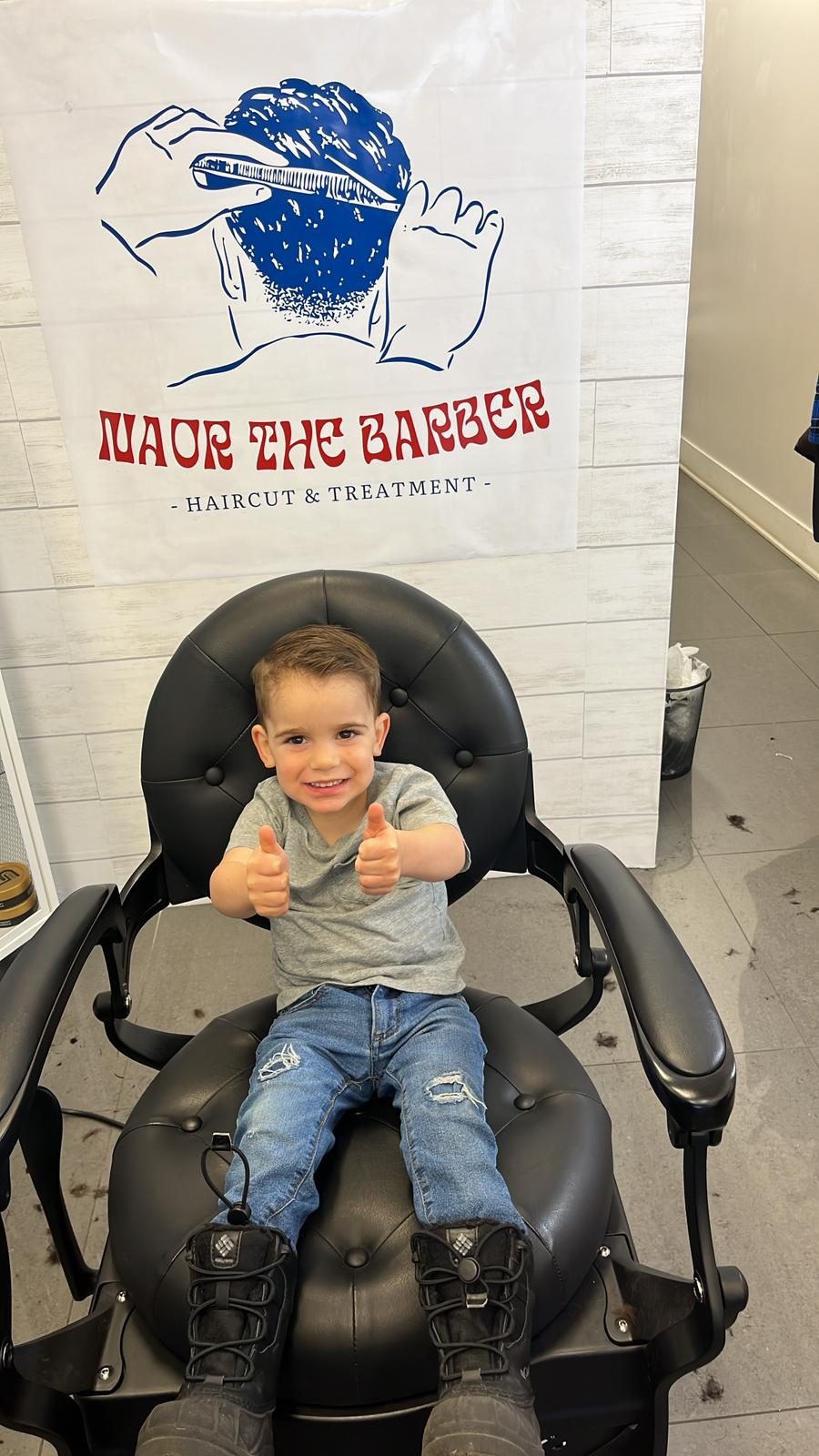 Jeune garçon qui fait deux pouces en l'air dans la chaise de barbier après sa coupe
